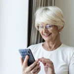Mujer mayor sonriente leyendo una conversación de WhatsApp en su móvil en casa