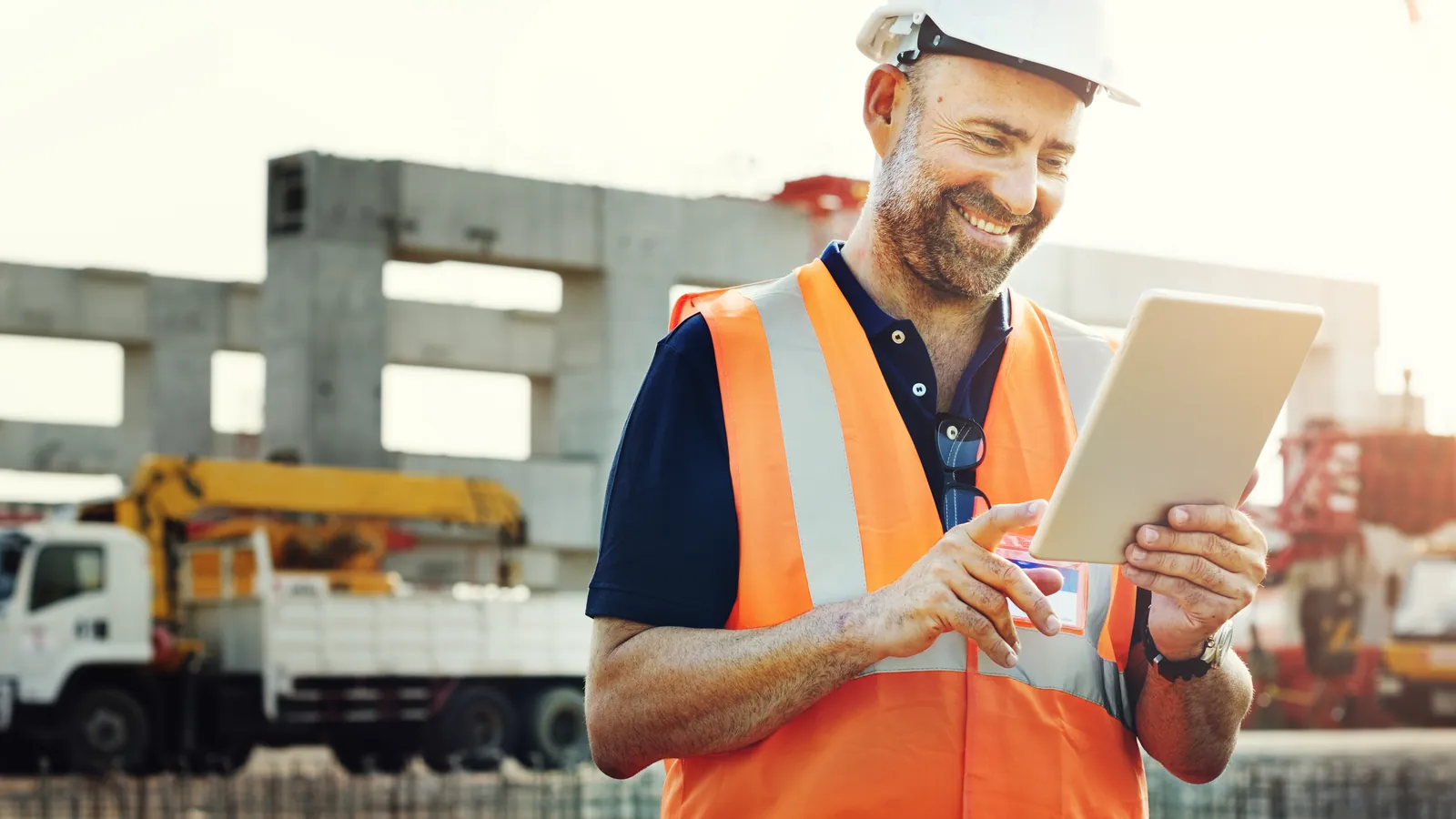 Técnico de prevención de riesgos laborales sonriente, con casco y chaleco, consultando una tableta en una obra