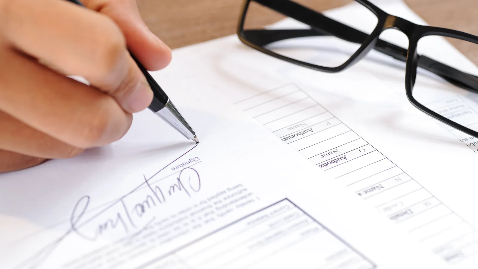 Primer plano de una mano firmando un documento junto a unas gafas