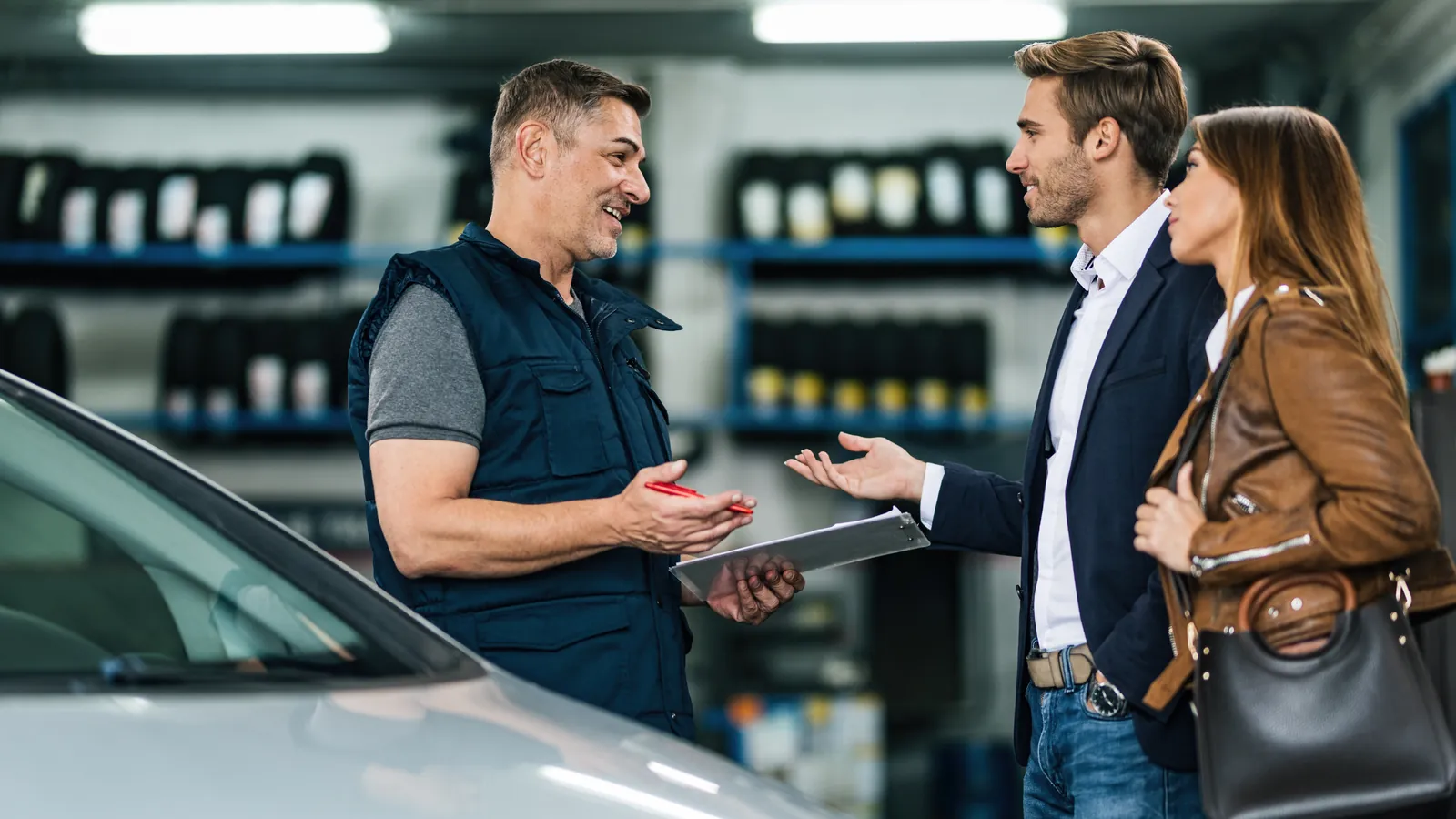 Perito de coches sonriente explicando la revisión a una pareja junto a su coche en el taller