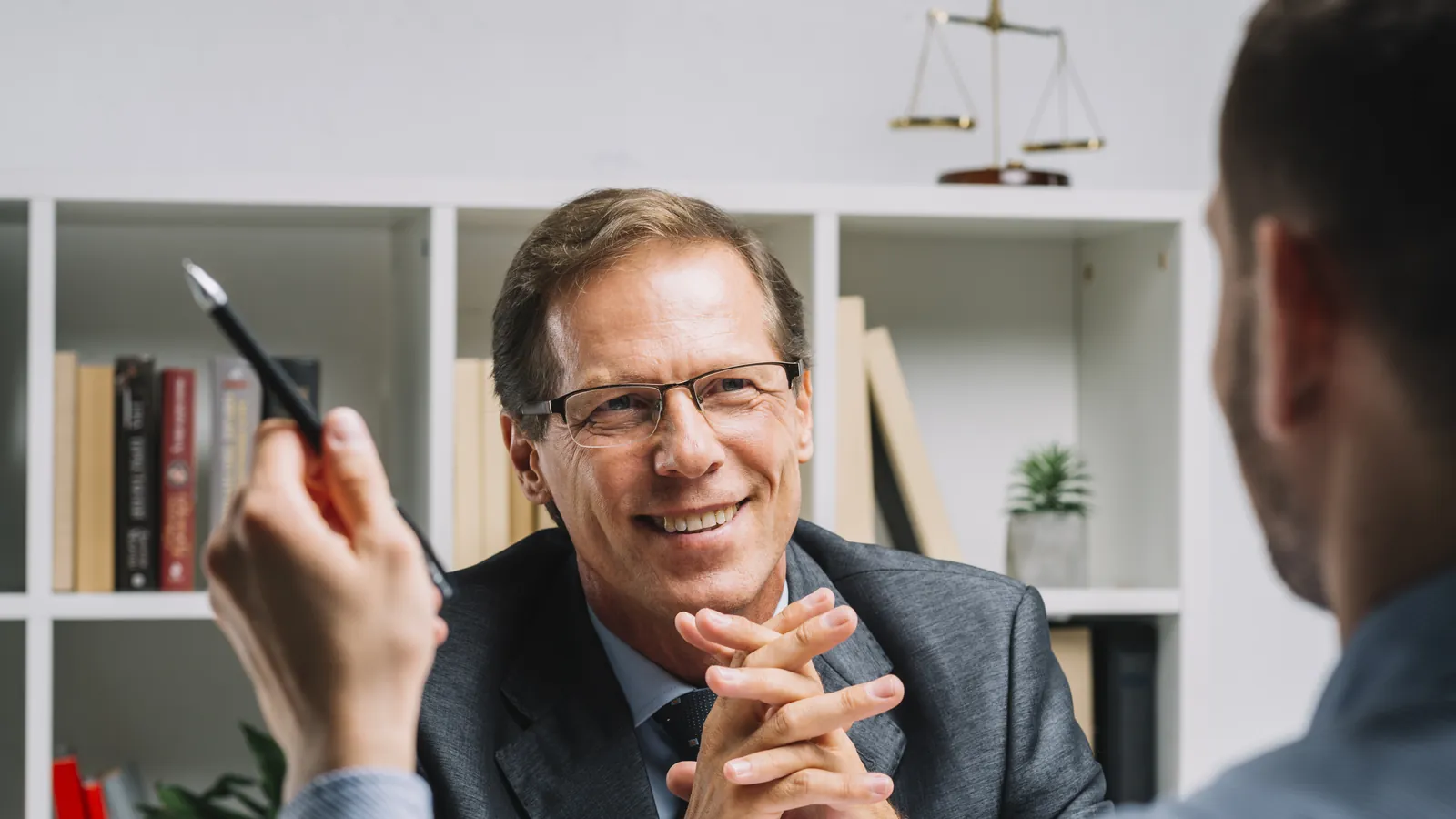 Perito judicial sonriente atendiendo a un cliente en su despacho, con una balanza de la justicia en la estantería