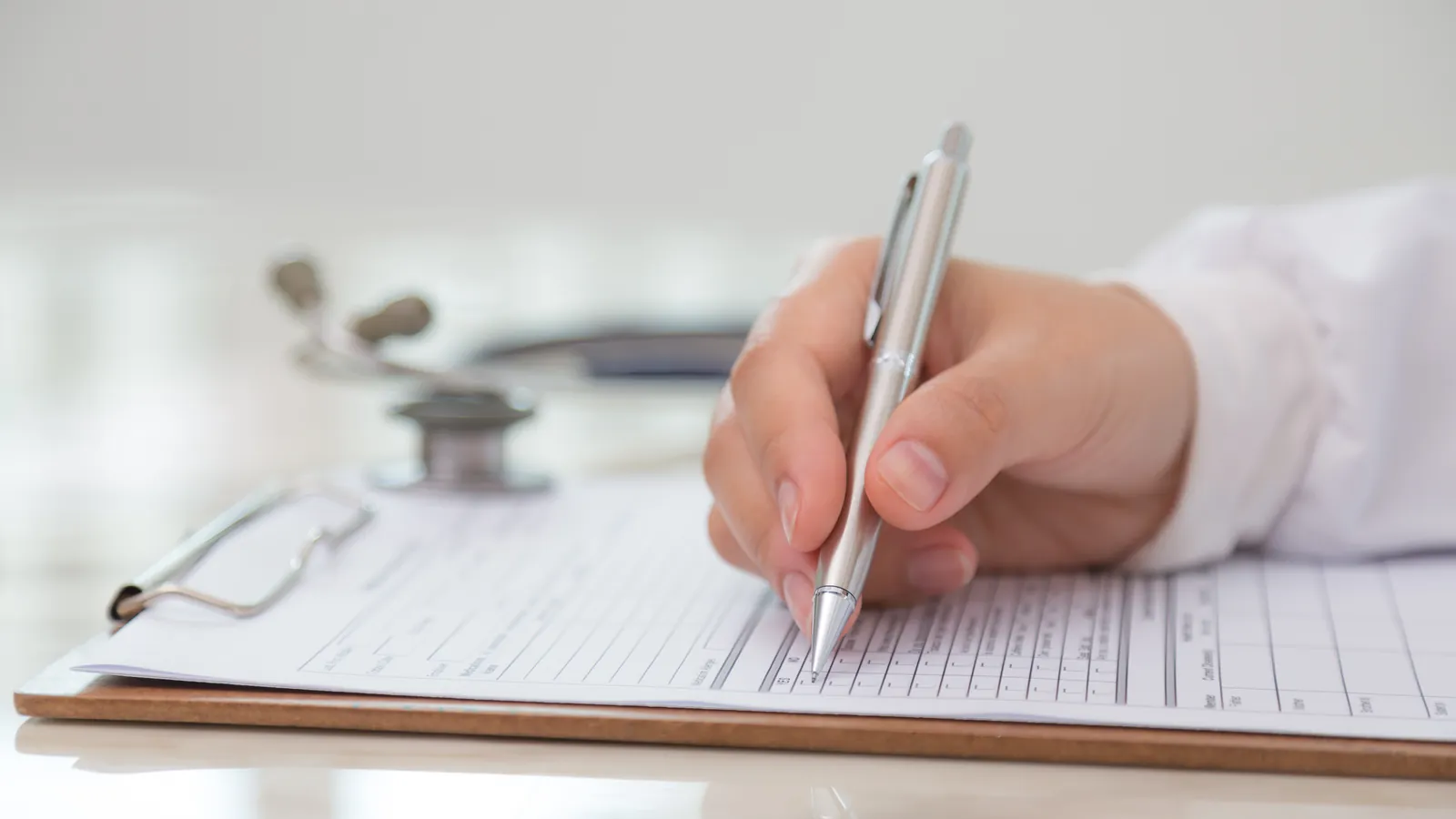 Mano de un médico rellenando un informe clínico sobre una tablilla, con el estetoscopio al fondo